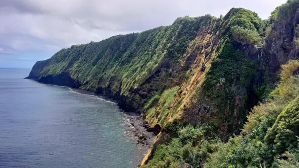 Falésias verdes a pique sobre um mar turquesa, com uma cascata a descer a rocha